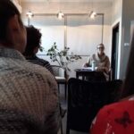Zdjęcie przedstawiające Adrianę Meller prowadzącą warsztaty lub spotkanie psychoanalityczne w kameralnej sali. Widoczni są uczestnicy siedzący na krzesłach oraz prowadząca przy stole.